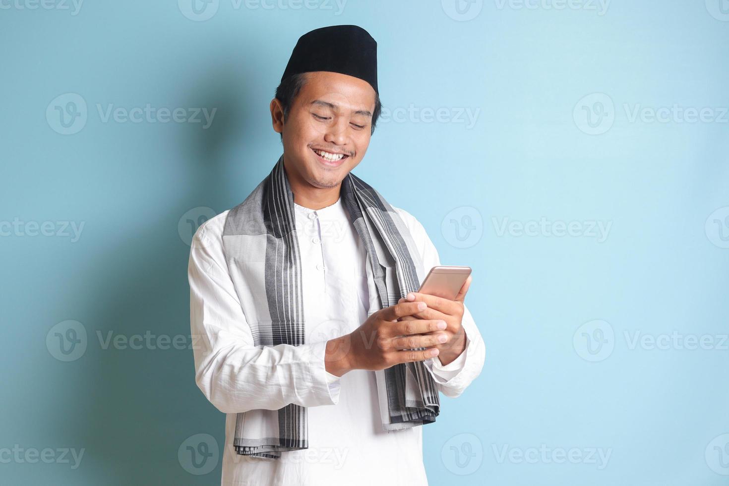 Portrait of young Asian muslim man holding and touching mobile phone with smiling expression on face. Isolated image on blue background photo