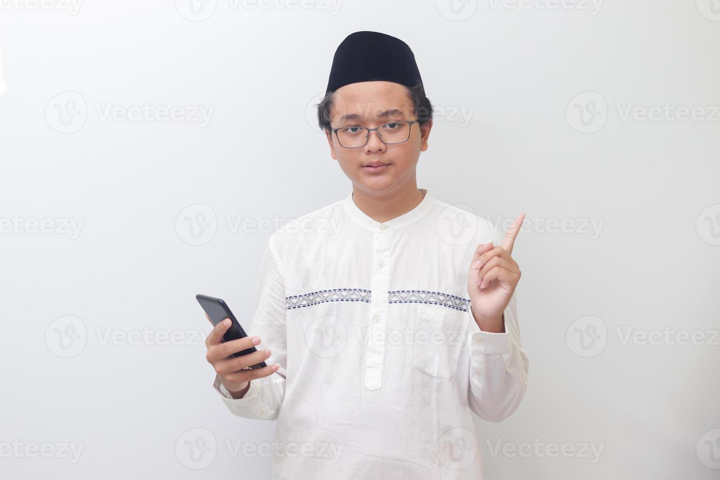 Portrait of young excited Asian muslim man pointing away with his finger while using mobile phone. Isolated image on white background photo
