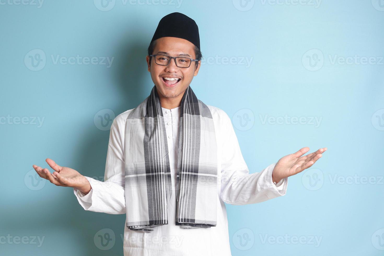 Portrait of Asian muslim man in white koko shirt with skullcap pulling hands towards camera, inviting someone to come inside, welcoming gesture. Isolated image on blue background photo