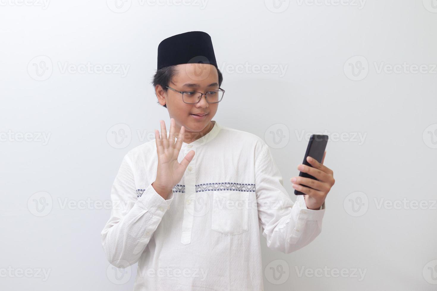 Portrait of smiling young Asian muslim man taking picture of himself or doing video call, saying hi and waving his hand. Isolated image on white background photo