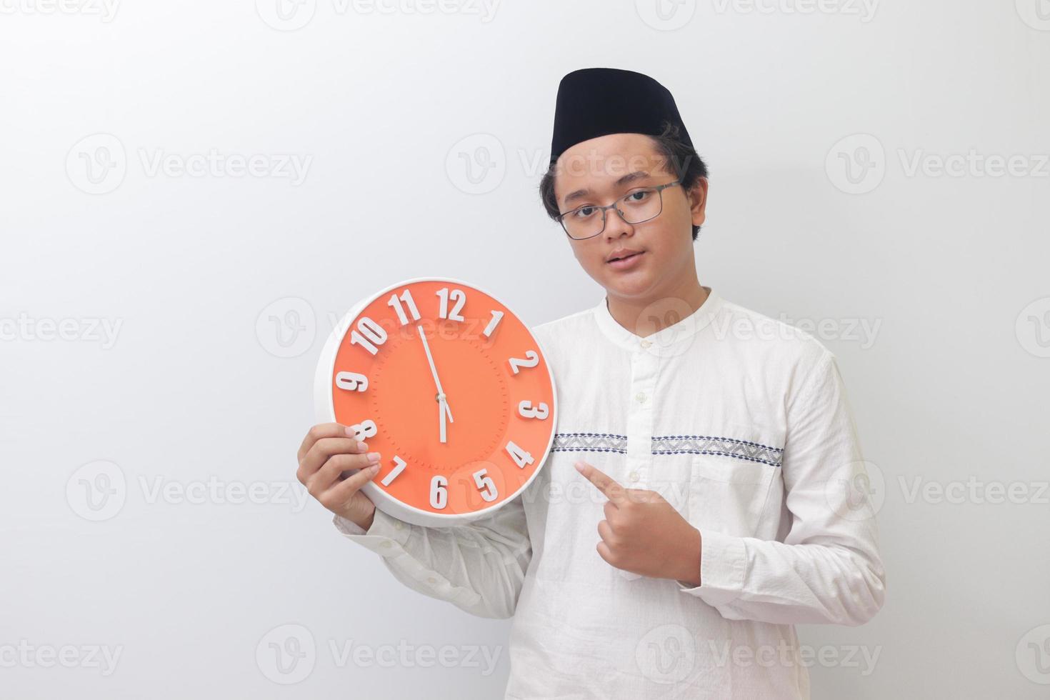 Portrait of young attractive Asian muslim man pointing and holding a clock that shows 6 pm. Isolated image on white background photo