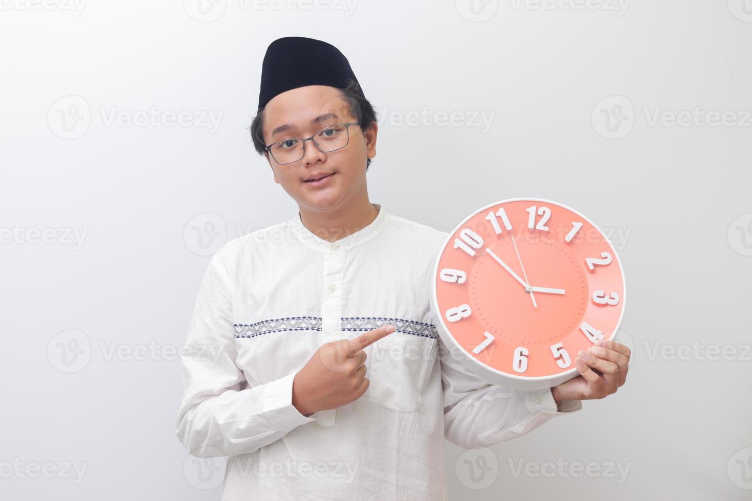 Portrait of young attractive Asian muslim man pointing and holding a clock that shows 3 am. Isolated image on white background photo