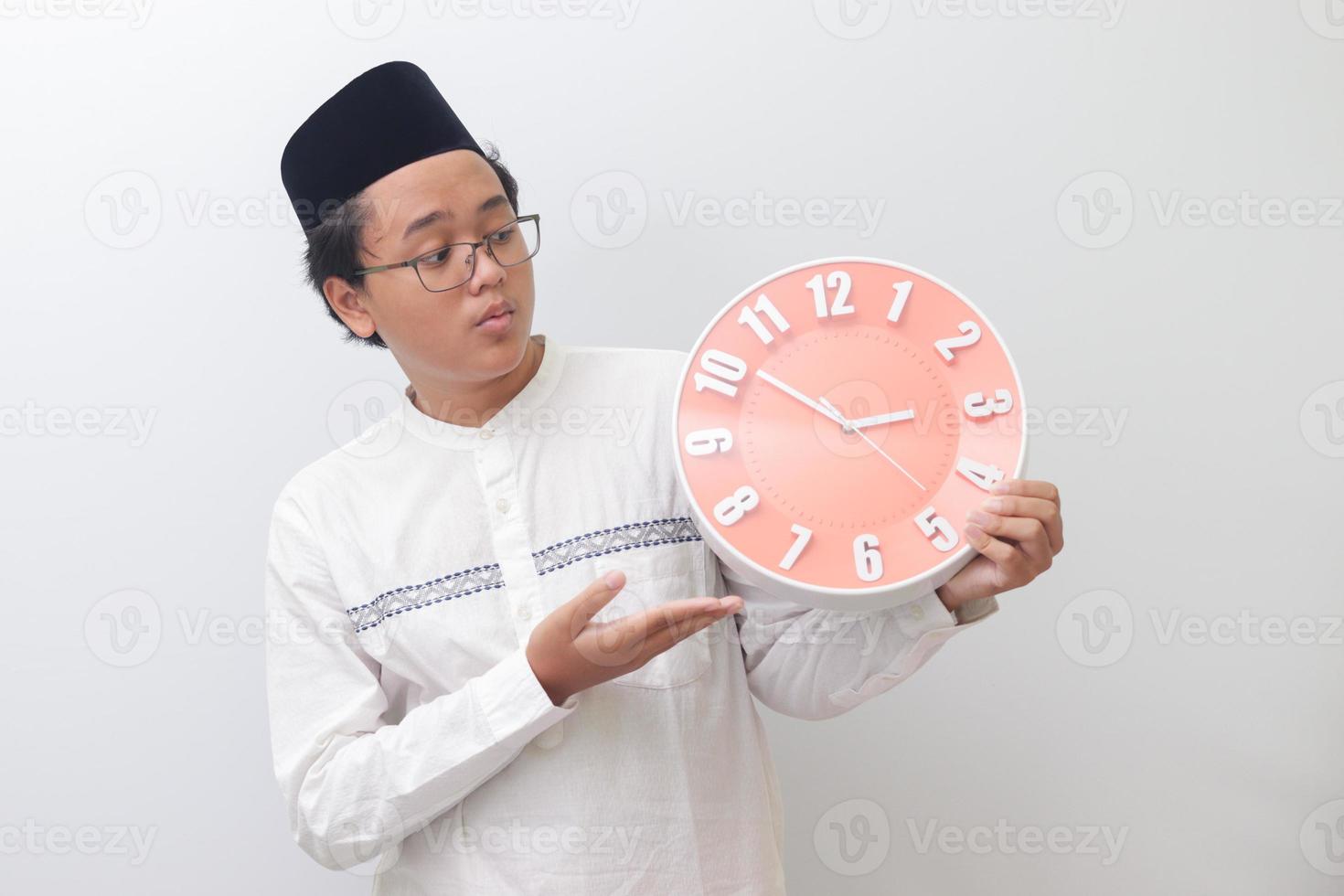 Portrait of young attractive Asian muslim man pointing and holding a clock that shows 3 am. Isolated image on white background photo