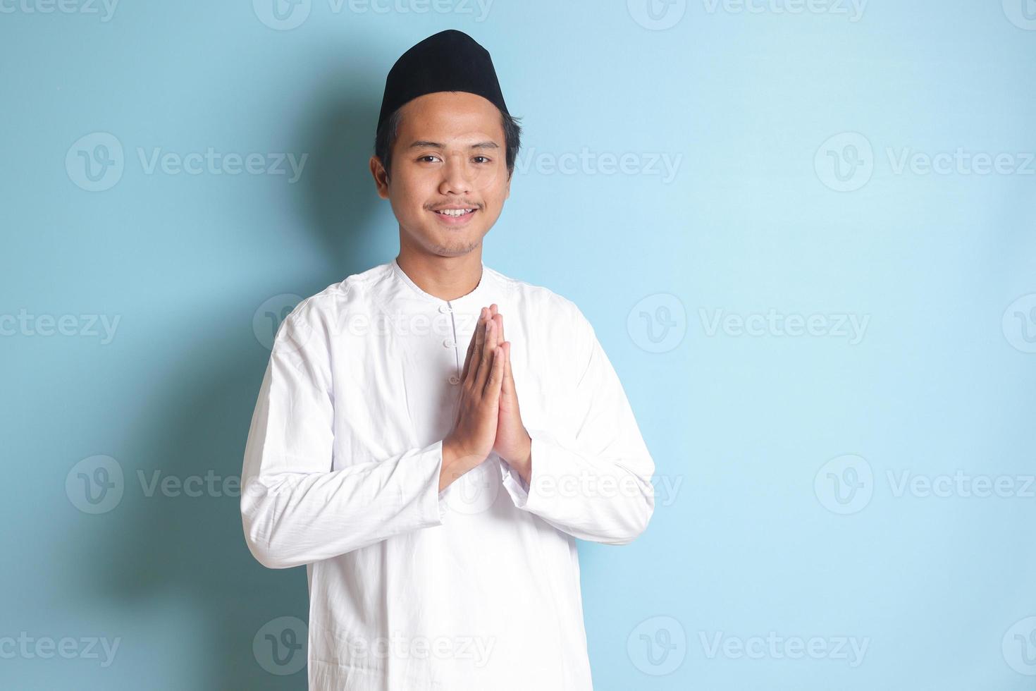 Portrait of Asian muslim man in white koko shirt with skullcap showing apologize and welcome hand gesture. Isolated image on blue background photo