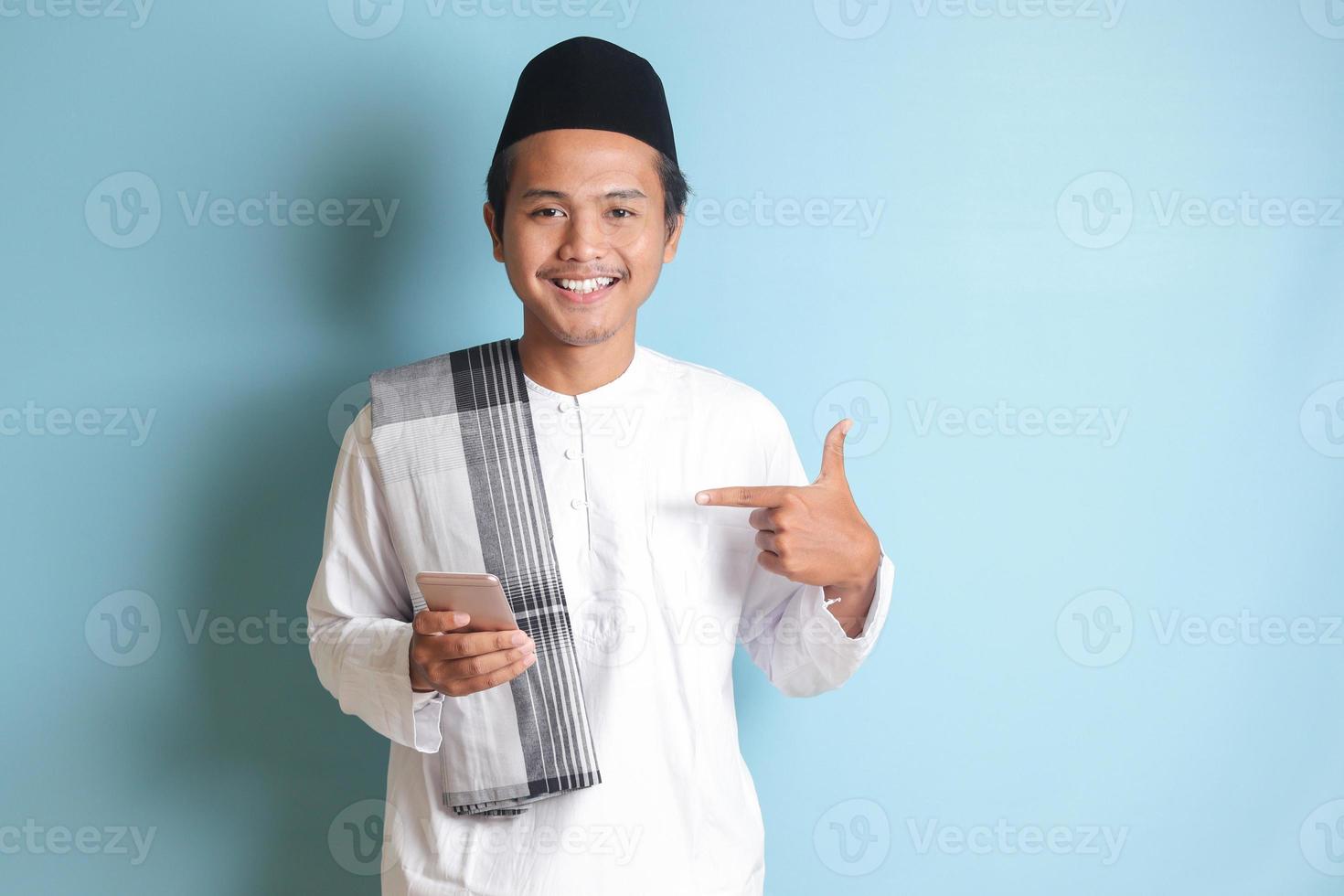 Portrait of young Asian muslim man holding mobile phone with smiling expression on face while pointing finger to the side. Isolated image on blue background photo