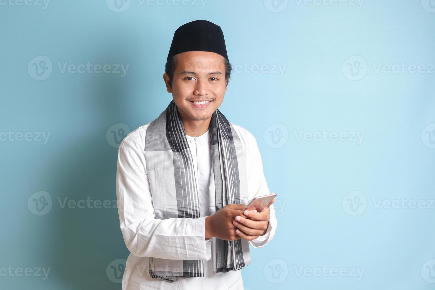 Portrait of young Asian muslim man holding and touching mobile phone with smiling expression on face. Isolated image on blue background photo