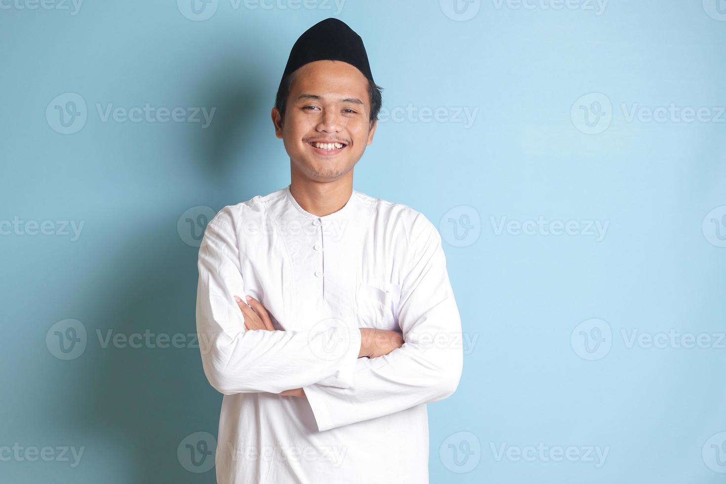 Portrait of religious Asian man in koko shirt or white muslim shirt and black cap, standing with crossed arms and looking at camera. Isolated image on blue background photo