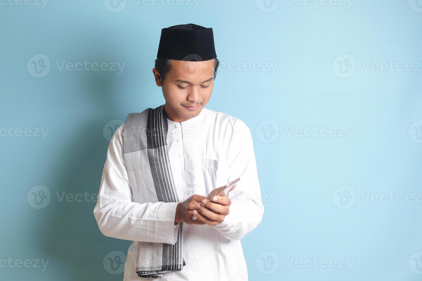 Portrait of young Asian muslim man holding and touching mobile phone with smiling expression on face. Isolated image on blue background photo