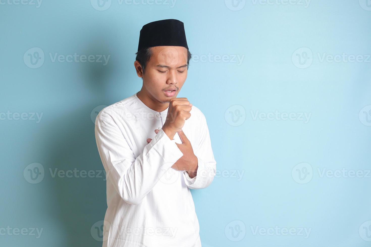 Portrait of attractive Asian muslim man in white shirt keeping hand palm covering his mouth while coughing with the other hand holding his chest. Isolated image on blue background photo