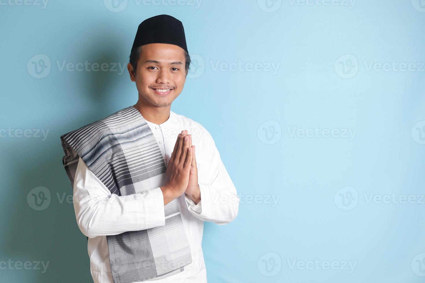 retrato de asiático musulmán hombre en blanco koko camisa con casquete demostración pedir disculpas y Bienvenido mano gesto. aislado imagen en azul antecedentes foto
