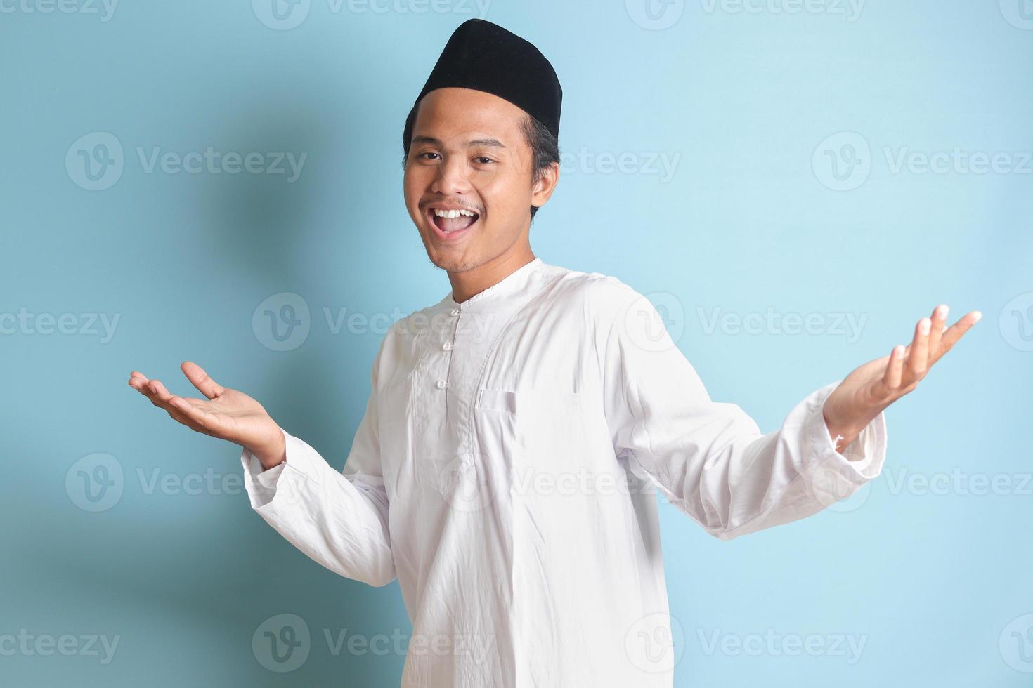 Portrait of Asian muslim man in white koko shirt with skullcap pulling hands towards camera, inviting someone to come inside, welcoming gesture. Isolated image on blue background photo