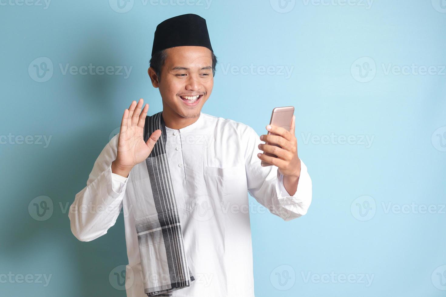 Portrait of Asian muslim man in white koko shirt with skullcap taking picture of himself or selfie, saying hi and waving his hand during video call. Isolated image on blue background photo