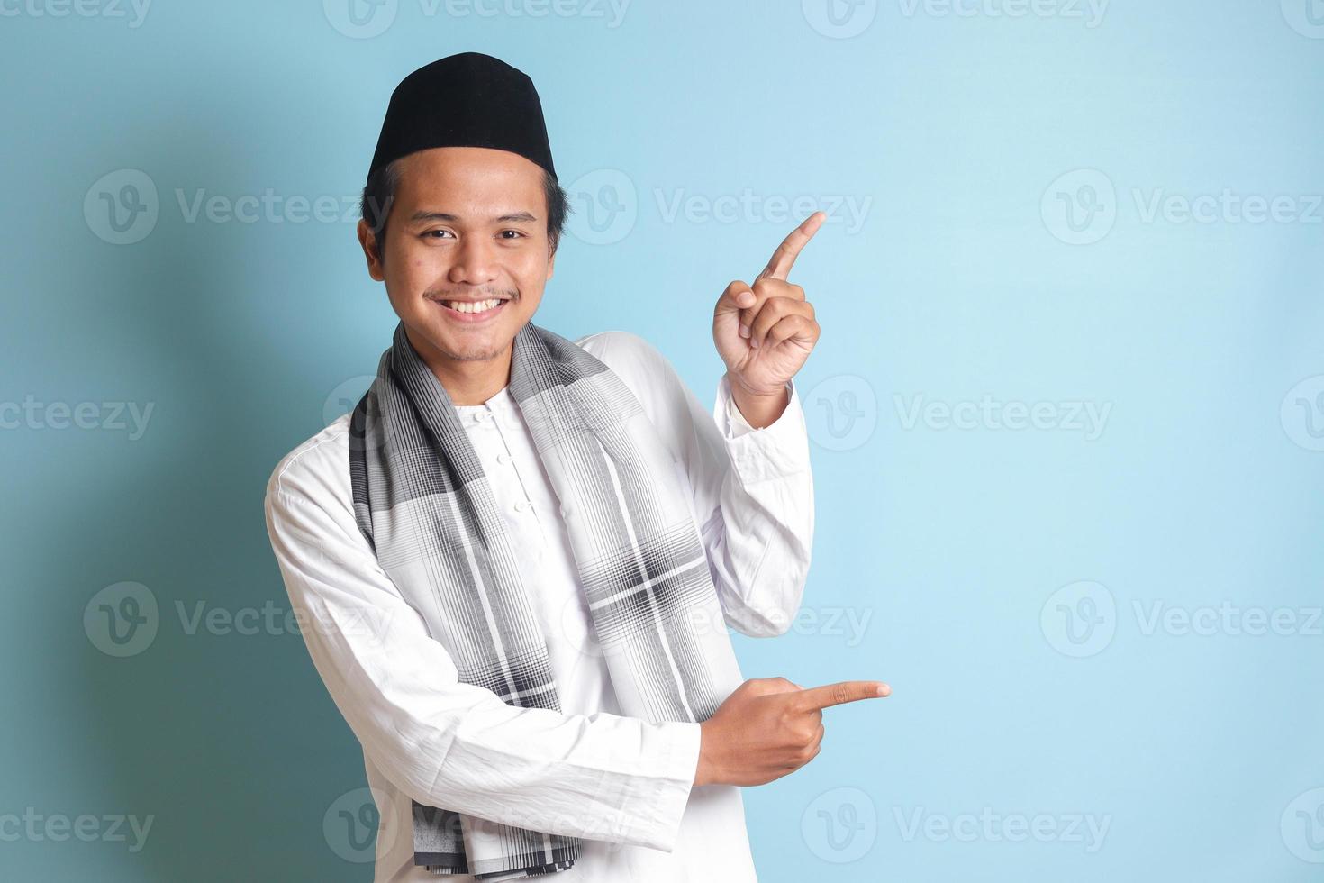 Portrait of young Asian muslim man showing product and pointing with his hand and finger to the side. Isolated image on blue background photo