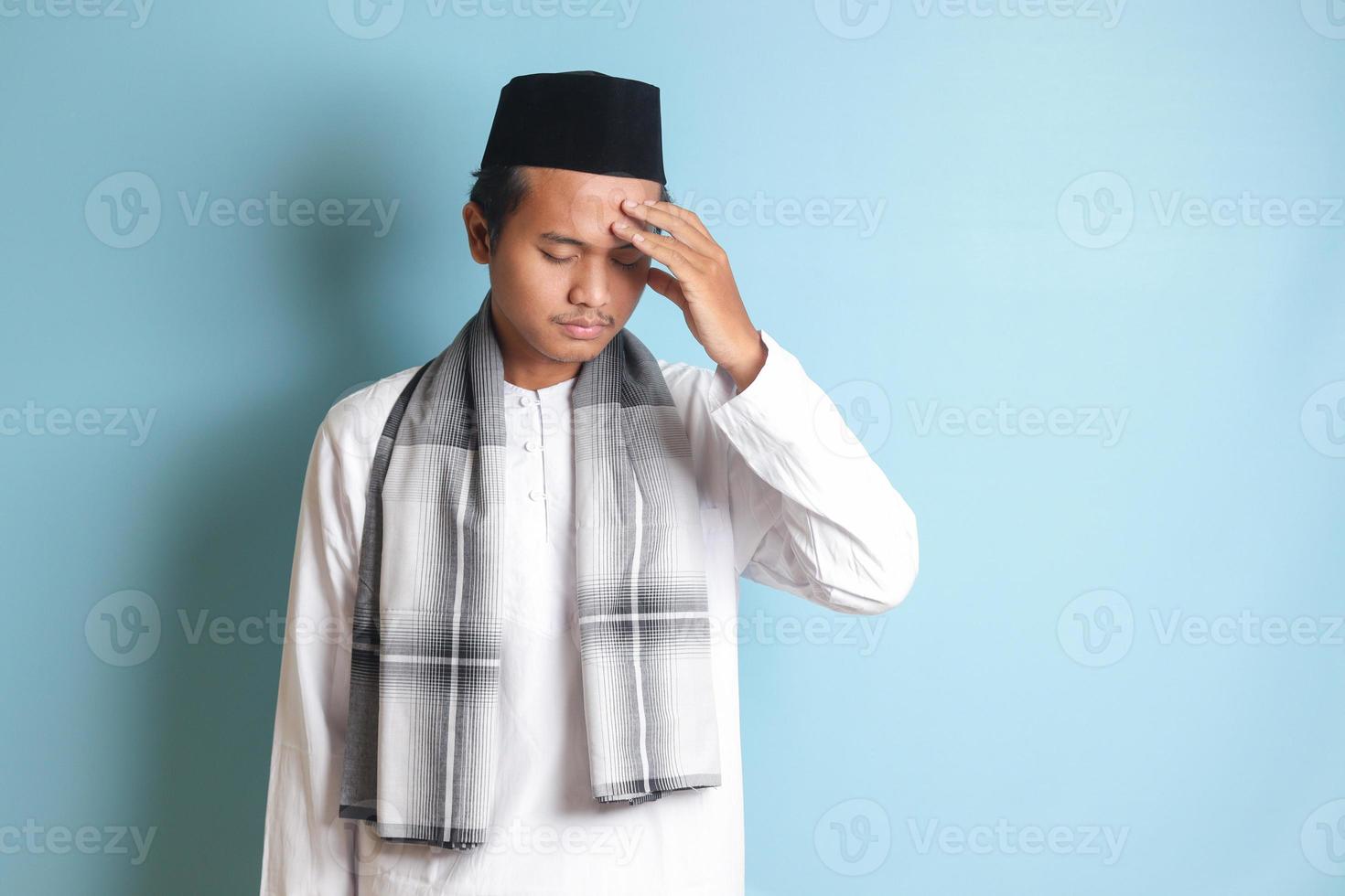 Portrait of attractive Asian muslim man in white shirt having a migraine, touching his temple. Headache disease concept. Isolated image on blue background photo