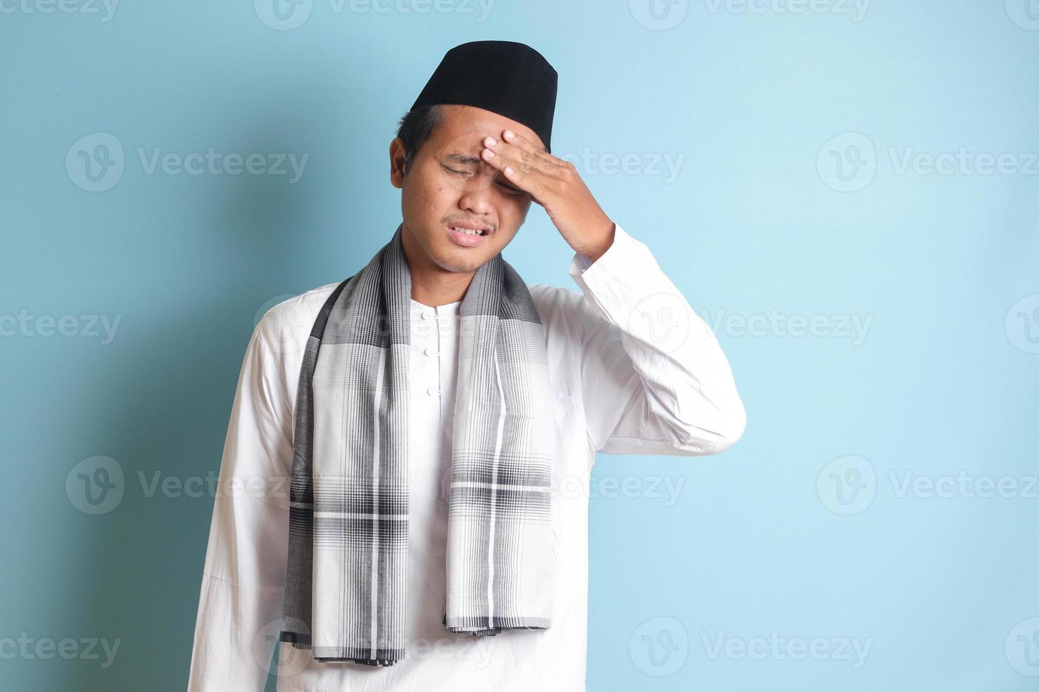 Portrait of attractive Asian muslim man in white shirt having a migraine, touching his temple. Headache disease concept. Isolated image on blue background photo