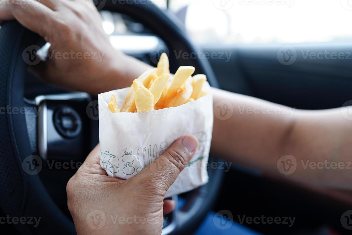 asiático mujer conductor sostener y comer francés papas fritas en auto, peligroso y riesgo un accidente. foto