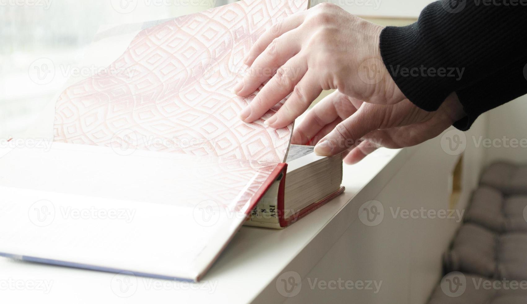 A person reading books near the window. Hands turns over book page. photo