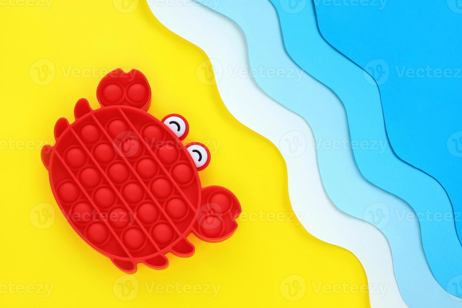 Red crab popular antistress sensory pop it toy on paper cut out seashore diagonal background, yellow sand and blue waves. Relaxation and fun on vacation for the whole family. Summer holidays concept. photo