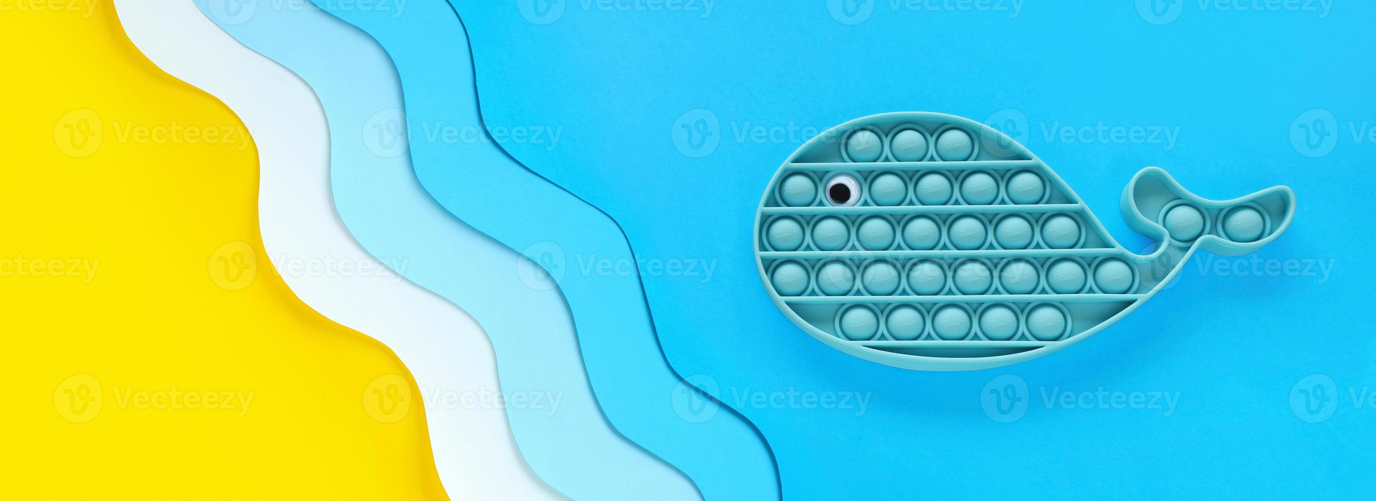 Blue whale popular antistress sensory silicone pop it toy on paper cut out seashore background, yellow sand and blue waves. For children and adults relaxation on vacation. Holidays summer long banner. photo