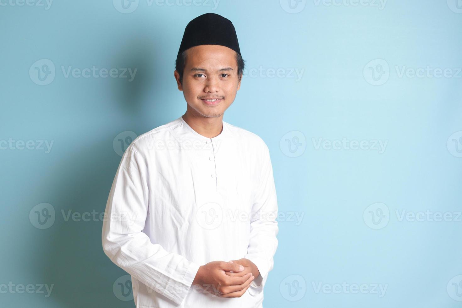 Portrait of Asian muslim man in white koko shirt feeling confident, smiling and looking at camera. Isolated in blue background photo