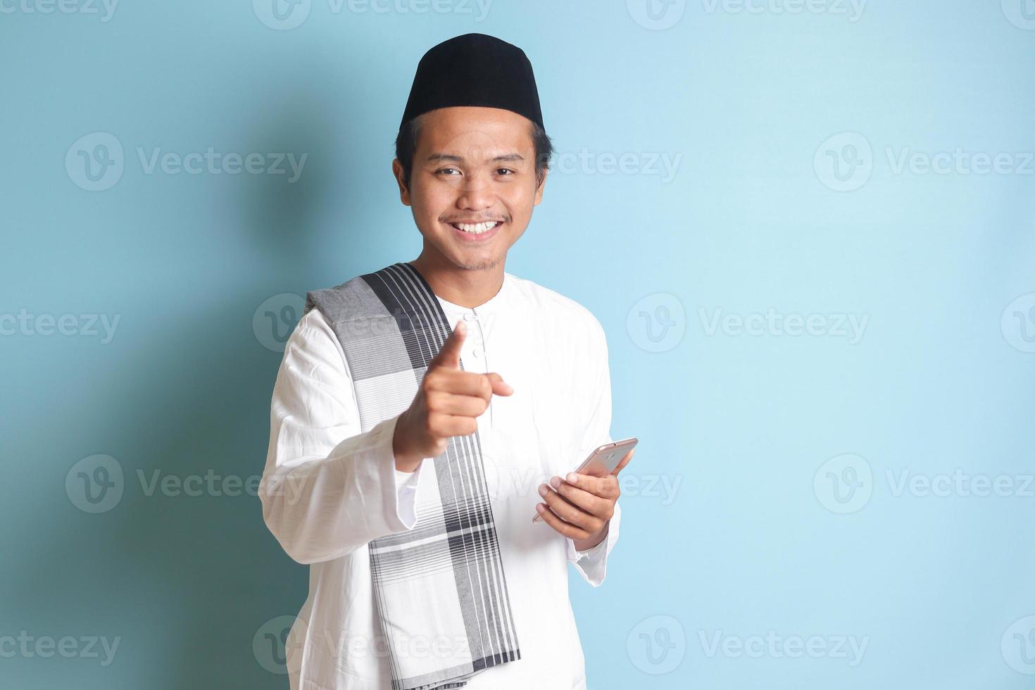 Portrait of young Asian muslim man holding mobile phone with smiling expression on face while pointing finger to the side. Isolated image on blue background photo