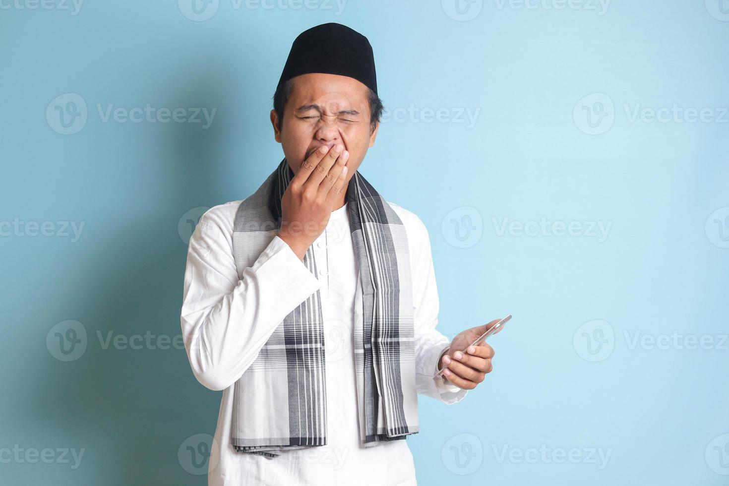 Portrait of young Asian muslim man holding mobile phone with while yawning with hand covering his mouth. Isolated image on blue background photo