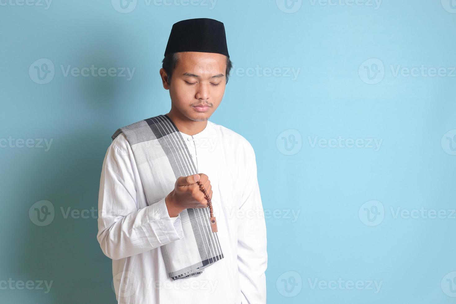 Portrait of Asian muslim man in white koko shirt with skullcap praying earnestly with his hands raised. Isolated image on blue background photo