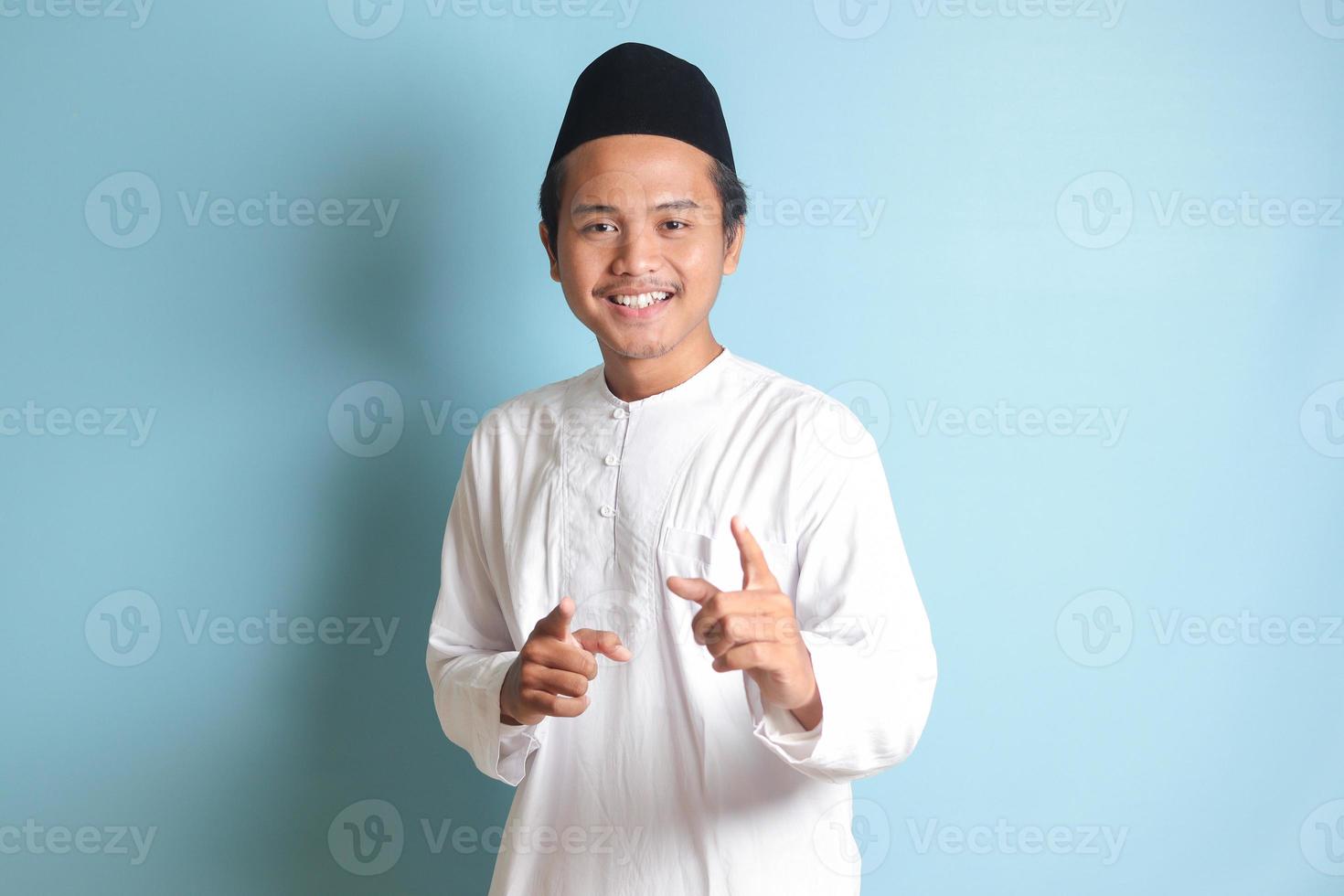 Portrait of young Asian muslim man showing product and pointing with his hand and finger to the side. Isolated image on blue background photo