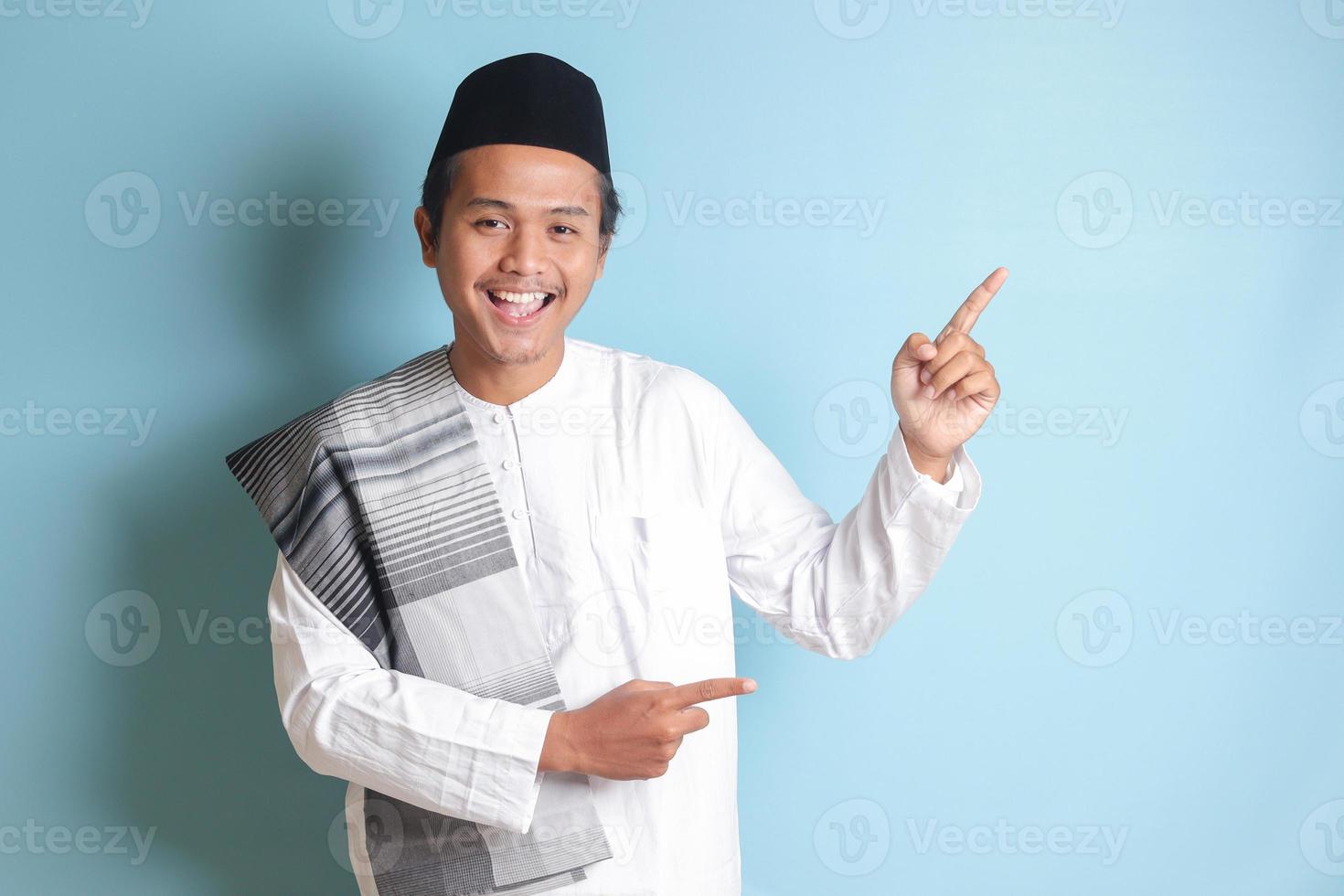 Portrait of young Asian muslim man showing product and pointing with his hand and finger to the side. Isolated image on blue background photo