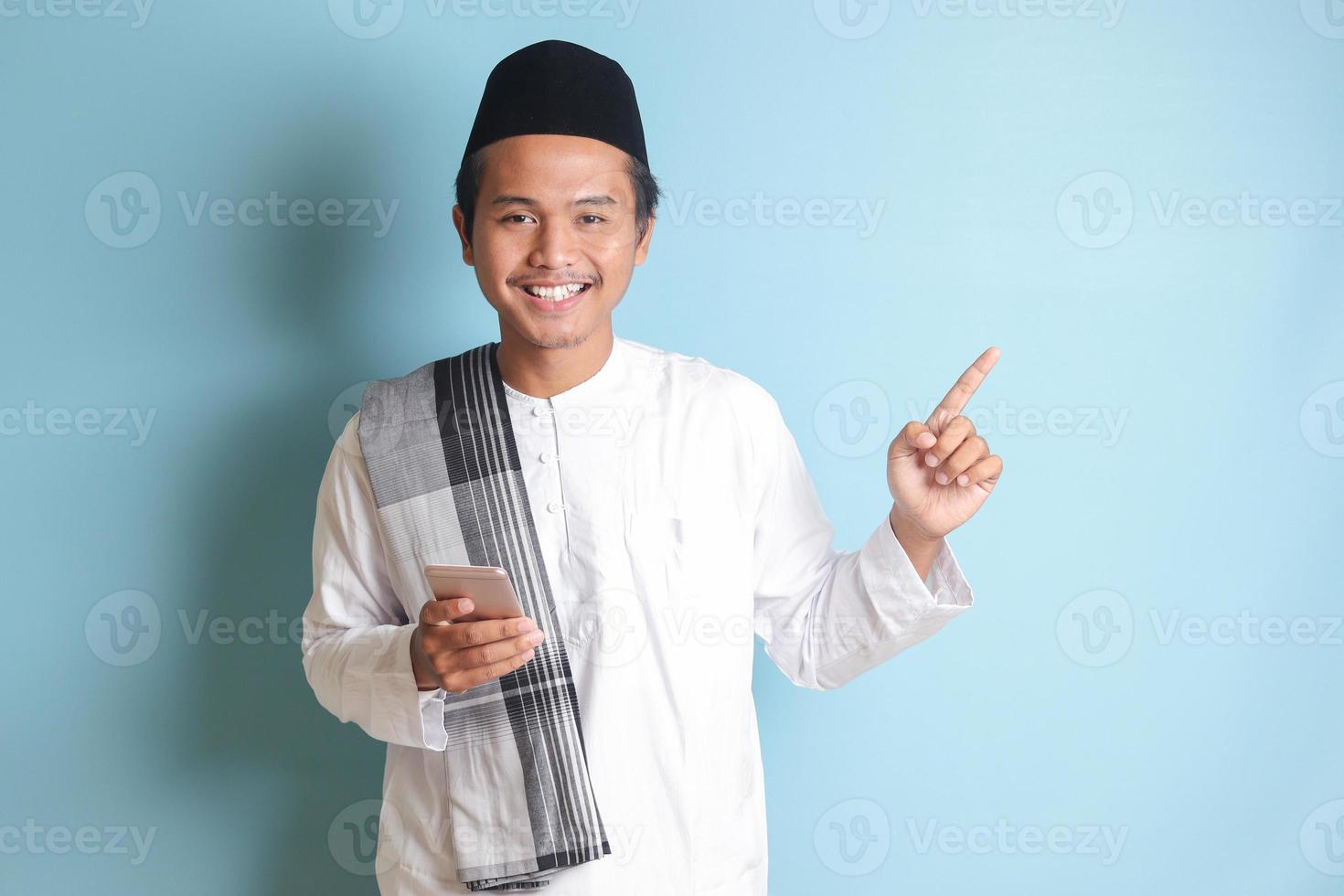 retrato de joven asiático musulmán hombre participación móvil teléfono con sonriente expresión en cara mientras señalando dedo a el lado. aislado imagen en azul antecedentes foto