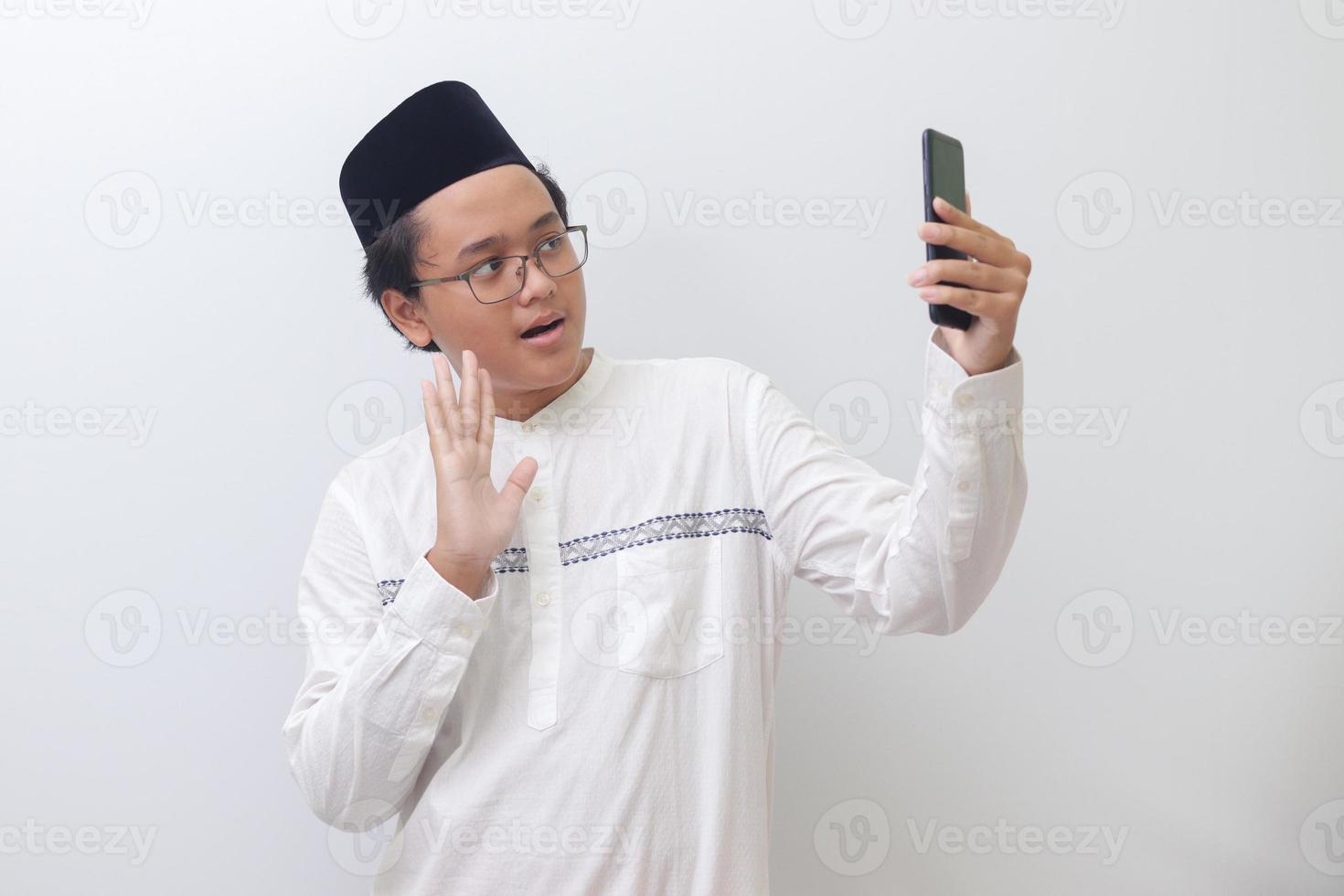 Portrait of smiling young Asian muslim man taking picture of himself or doing video call, saying hi and waving his hand. Isolated image on white background photo