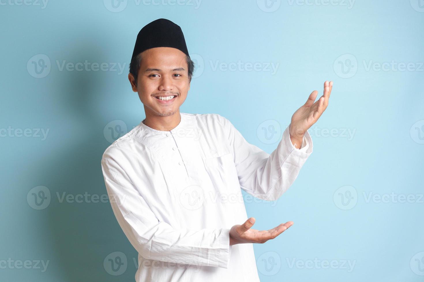 Portrait of young Asian muslim man showing product and pointing with his hand and finger to the side. Isolated image on blue background photo