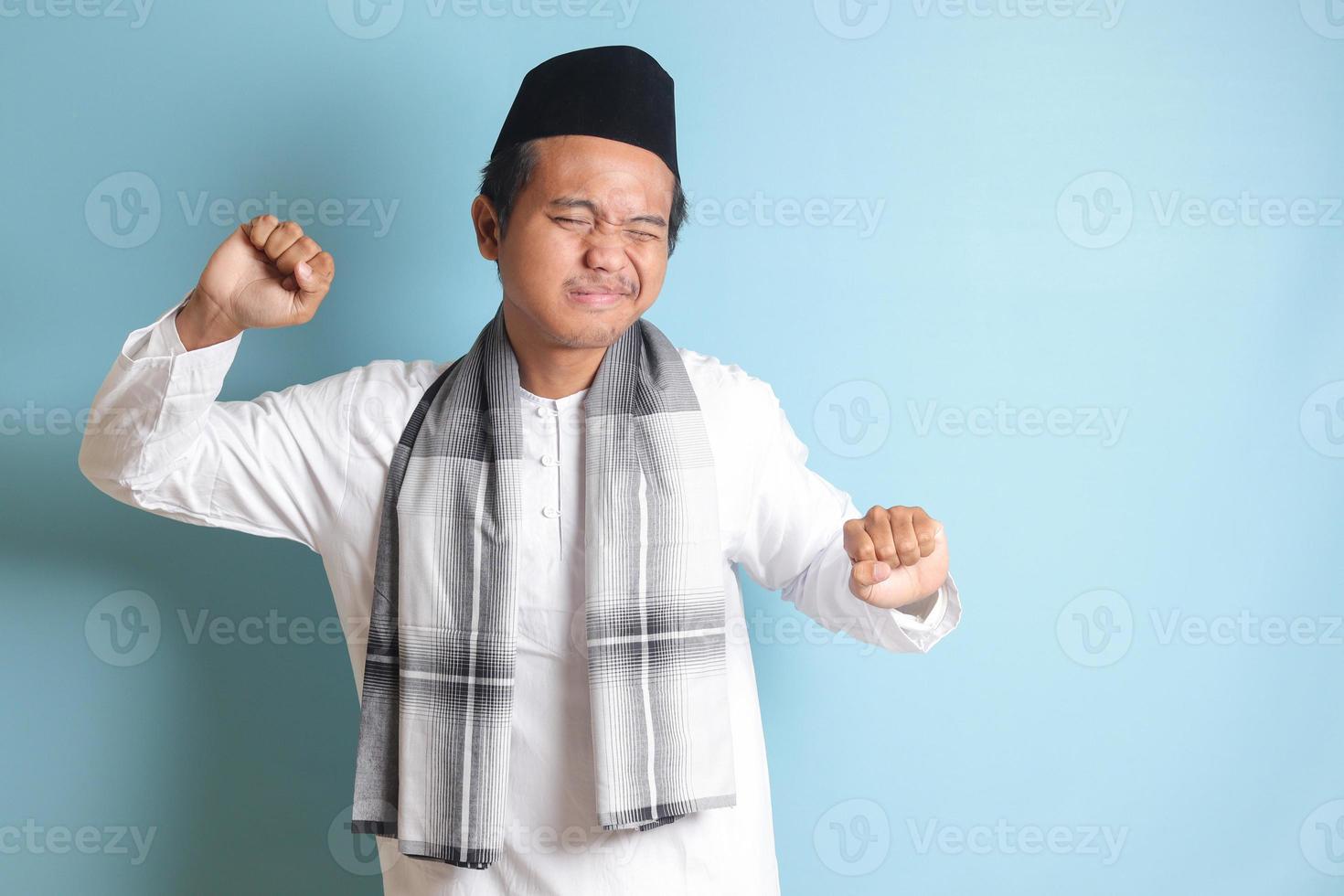 Portrait of attractive Asian muslim man in white shirt stretching his hands and body after waking up. Isolated image on blue background photo