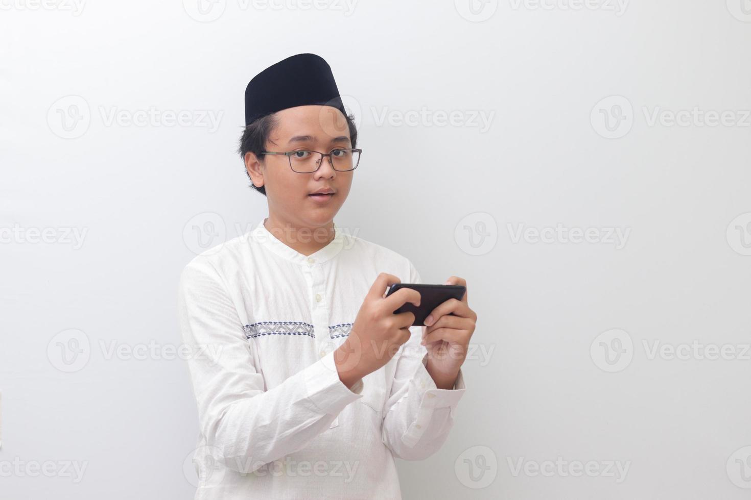 retrato de joven asiático musulmán hombre levantamiento su puño, celebrando victorioso juego o consiguiendo bueno Noticias en su móvil teléfono. aislado imagen en blanco antecedentes foto