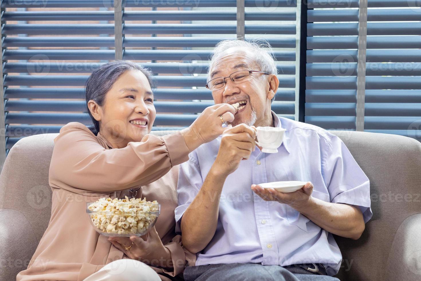 mayor asiático Pareja es sonriente mientras sentado en el sofá sofá y teniendo divertido acecho televisión con palomitas de maiz como bocadillo para bueno mental salud y longevidad concepto foto