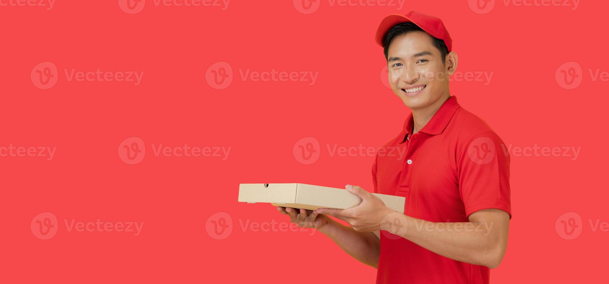 The smiling pizza delivery man stands on a red background holding the pizza box and wearing a red cap and a blank t-shirt uniform. photo