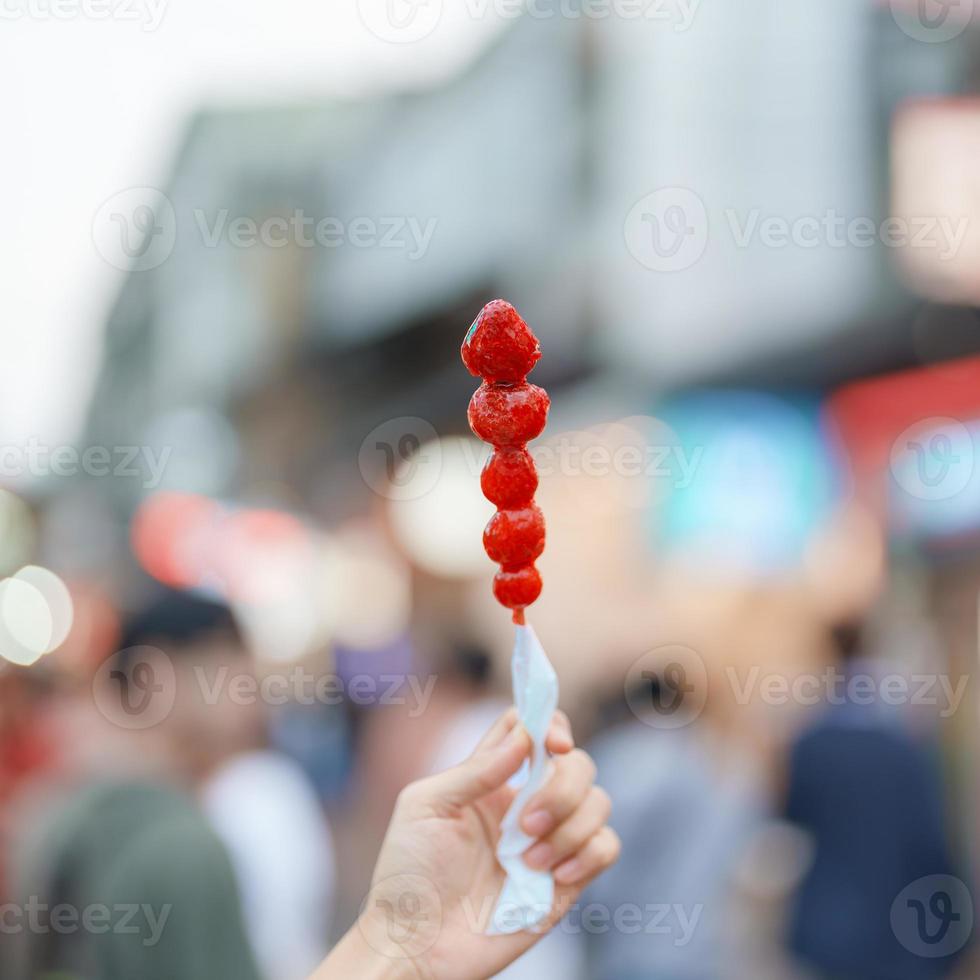 woman hand holding caramel coated strawberry skewer at night market. Street Food and travel concept photo