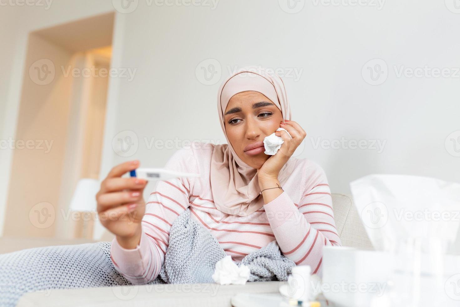 enfermedad, estacional virus problema concepto. Arábica mujer siendo enfermo teniendo gripe acostado en cama mirando a temperatura en termómetro. enfermo mujer con hijab acostado en cama con alto fiebre. foto