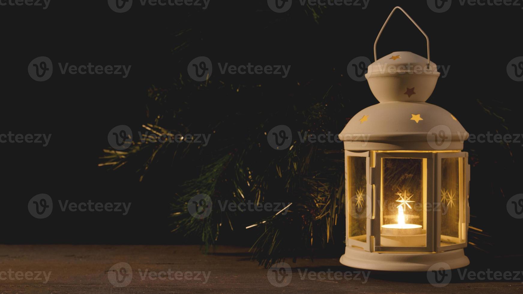 White candle lantern with burning candle. Blurred spruce brunch on the dark background. Retro style picture. photo