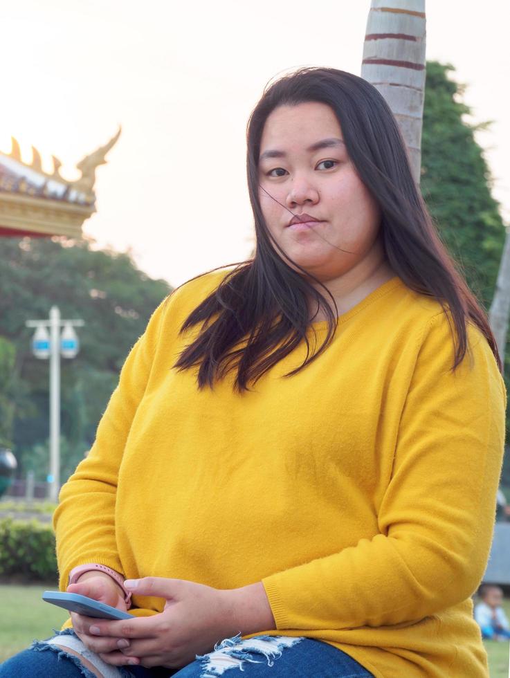 retrato grasa asiático mujer largo negro pelo vistiendo amarillo camisa son utilizando móvil teléfono o teléfono inteligente trabajo con grave y grave cara y expresión dentro parque en noche horas puesta de sol día foto