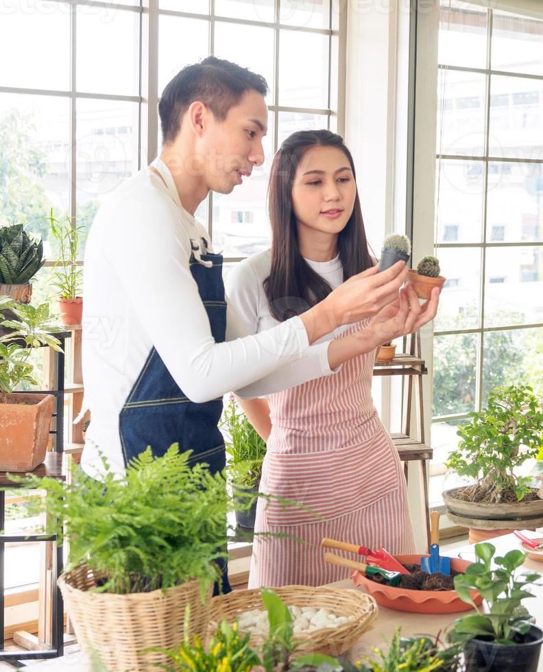 Gardener young Asian man woman family two person stand smiling looking hand holding help decorate the table tree leaf green in calm work shop home plant window wall. hobby job happy and care concept photo