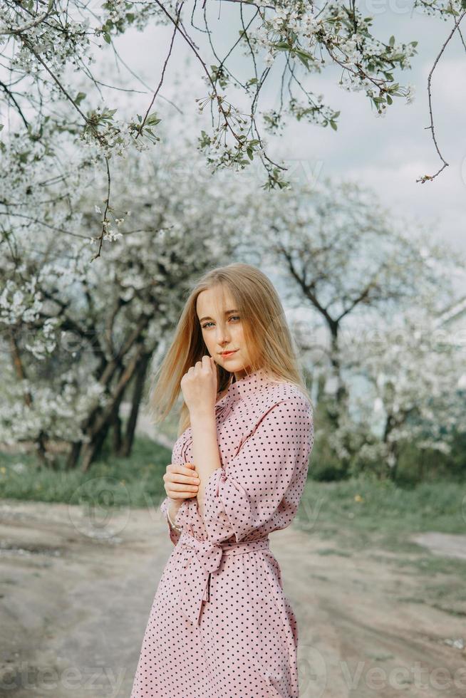 Blonde girl on a spring walk in the garden with cherry blossoms. Female portrait, close-up. A girl in a pink polka dot dress. photo