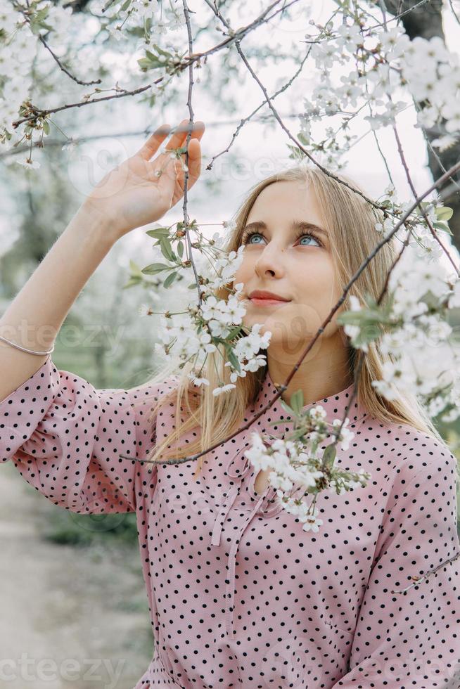 Blonde girl on a spring walk in the garden with cherry blossoms. Female portrait, close-up. A girl in a pink polka dot dress. photo