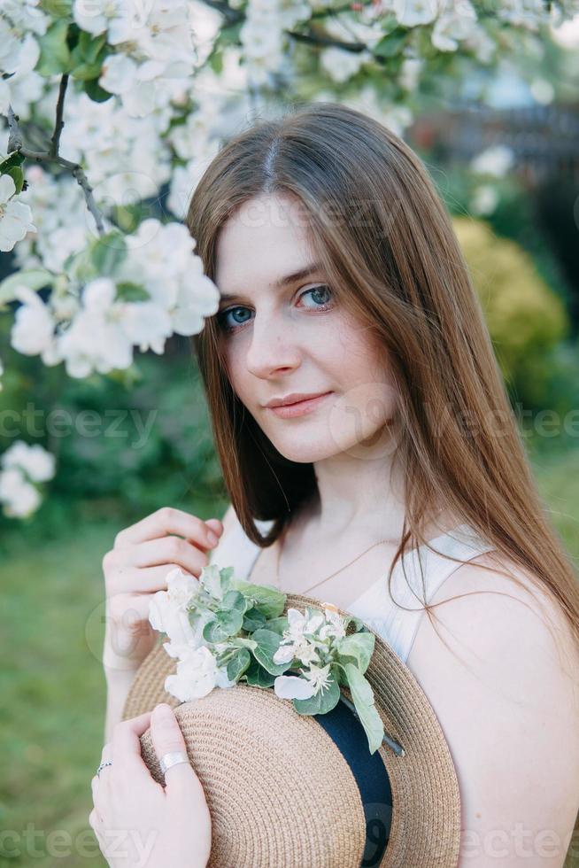Beautiful young girl in white dress and hat in blooming Apple orchard. Blooming Apple trees with white flowers. photo