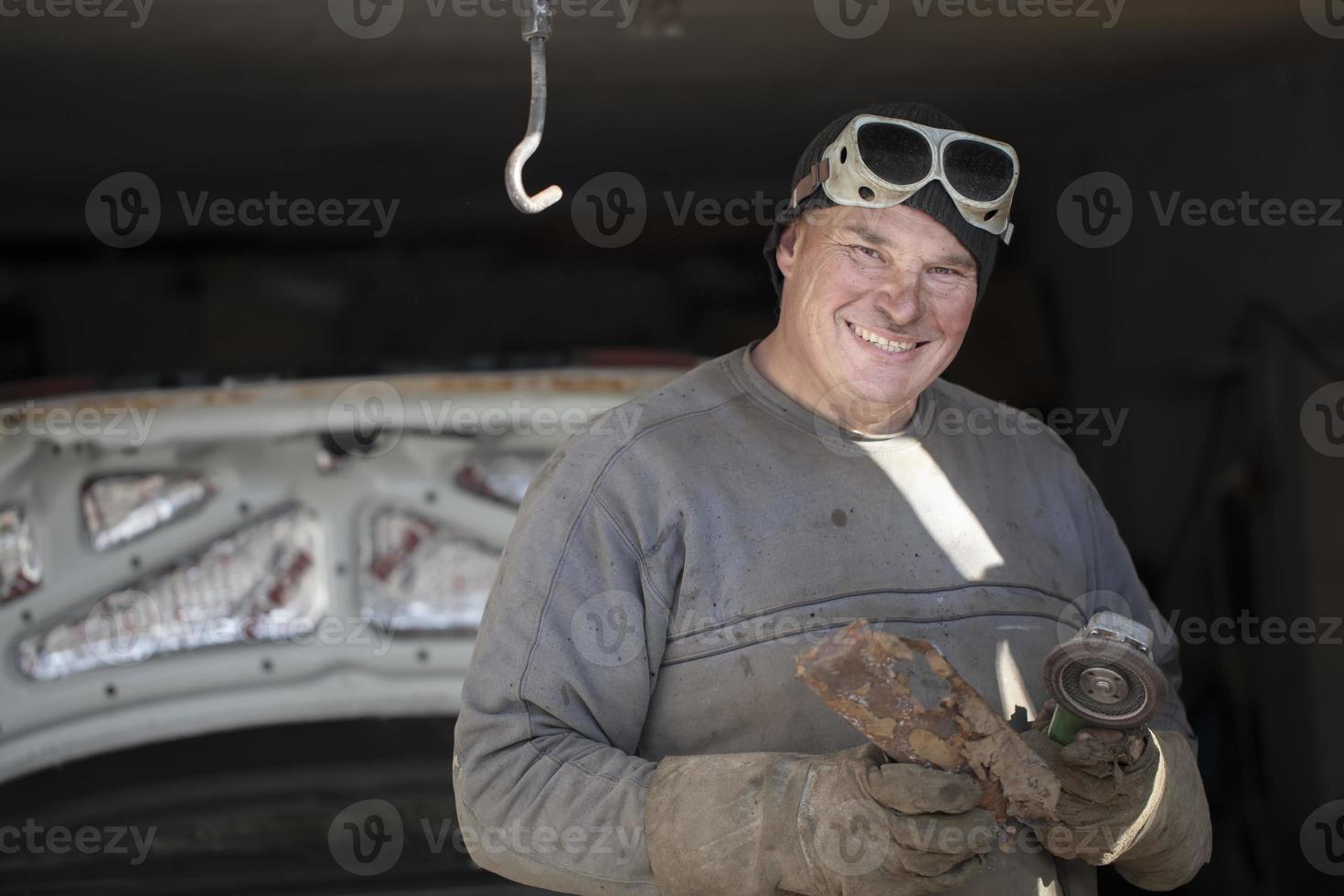 Cheerful male auto mechanic in dirty clothes in the garage holds a grinder. photo