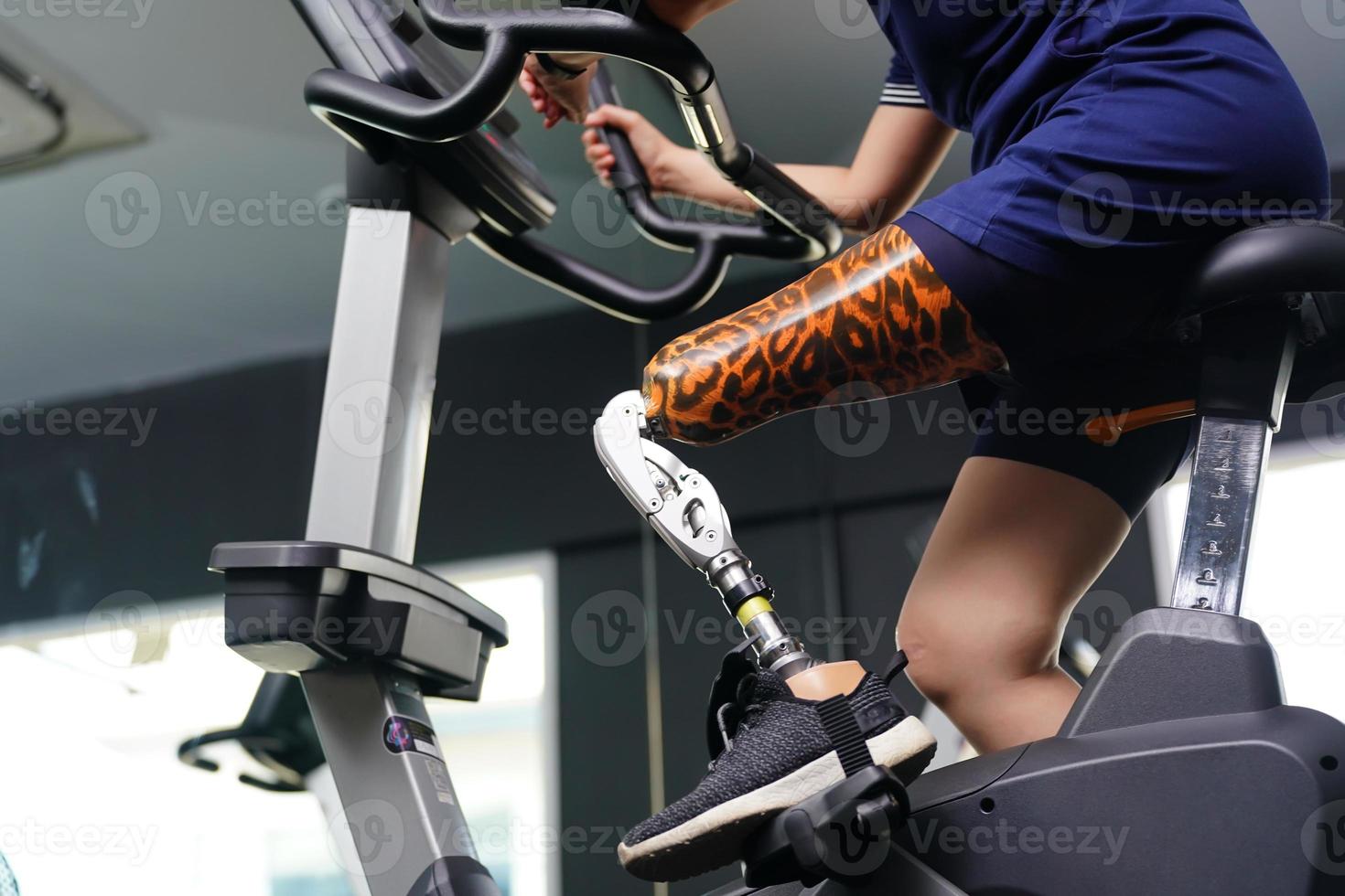 Young female with one prosthetic leg with exercising with a spinning bike in the gym to practice joint use with the other normal leg to be flexible, Concept of living a woman's life with a prosthetic photo