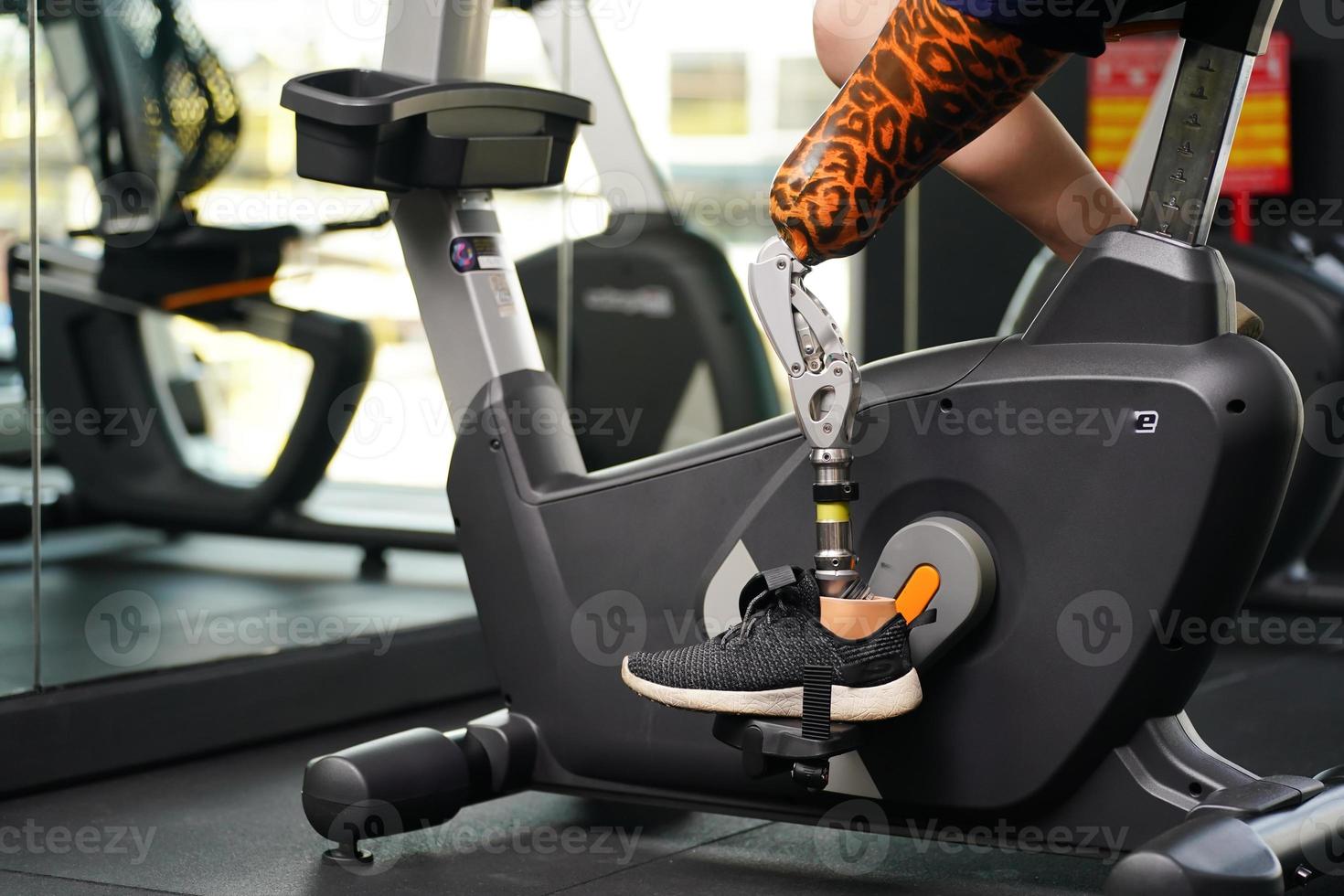 Young female with one prosthetic leg with exercising with a spinning bike in the gym to practice joint use with the other normal leg to be flexible, Concept of living a woman's life with a prosthetic photo