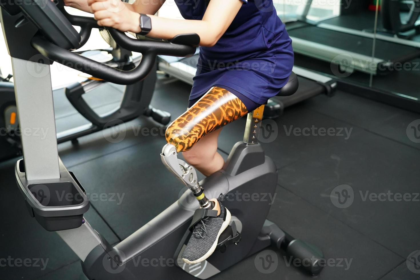 Young female with one prosthetic leg with exercising with a spinning bike in the gym to practice joint use with the other normal leg to be flexible, Concept of living a woman's life with a prosthetic photo