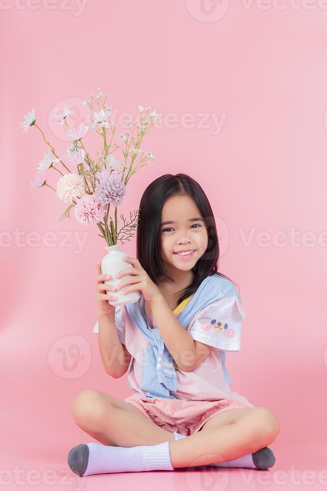 A little girl in a white dress is sitting near the window and holding a rose. A happy child at the window with a flower. photo