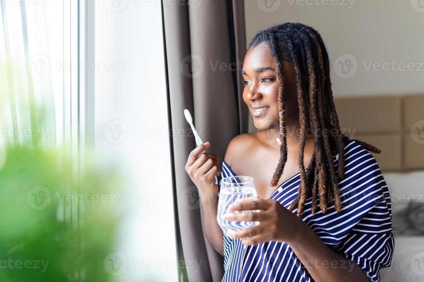 joven africano mujer cepillado dientes con cepillo de dientes, participación vaso de agua y mirando en ventana foto